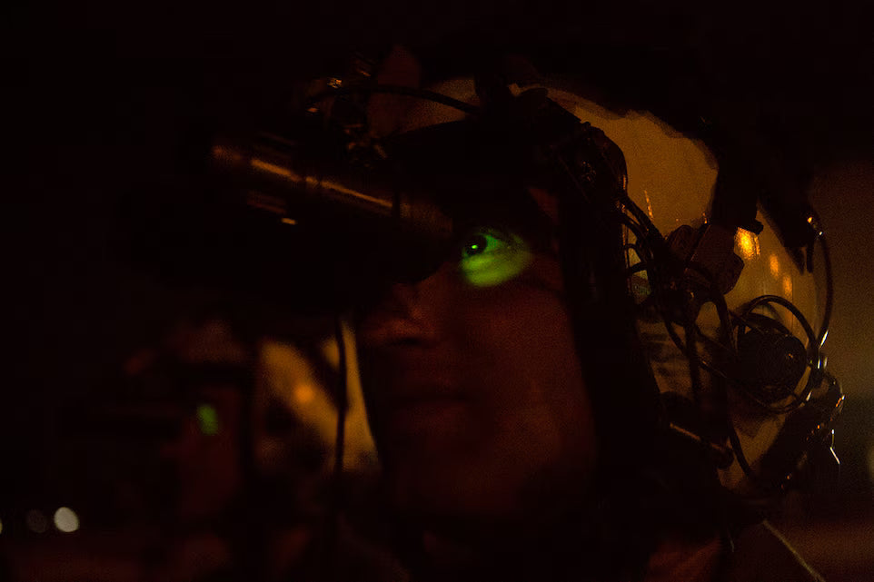 an image of a military personnel using a night vision device on operations where the image is mostly dark and there is an orange or red light glowing behind the person and it shows a glimpse of the helmet and side of his face