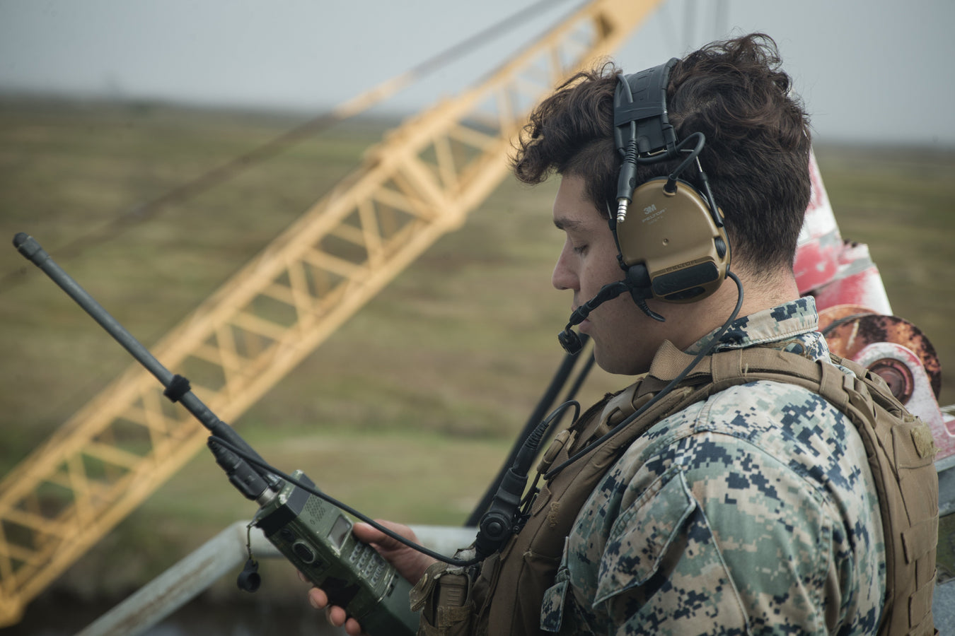 an image of a military personnel who is out on the field and looks to be on duty and he is operating a handheld radio along with a peltor comtac headset that he also seems to be using with the radio and is worn over his head