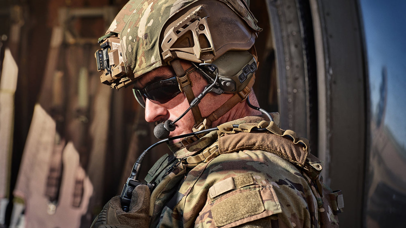 an image of a soldier in the field that is wearing a military uniform with matching colored military helmet where he is also using a 3m peltor headset and utilized the ptt feature as he speaks into the microphone