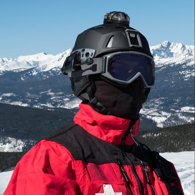A search and rescue operator stands before snow-capped mountains, wearing a black team wendy m-216 ski helmet equipped with a top-mounted strobe light and protective goggles.