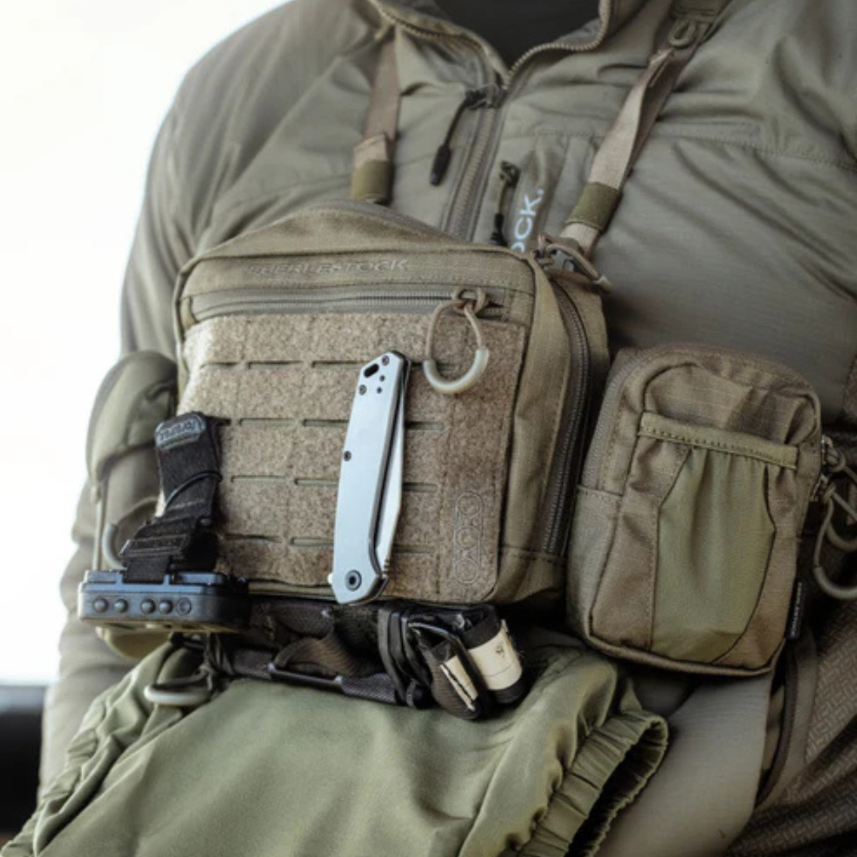 A front-angled view of a person wearing a modular chest rig in olive green, featuring a silver folding knife mounted on the front and a zippered utility pouch attached to the right side for extra storage.