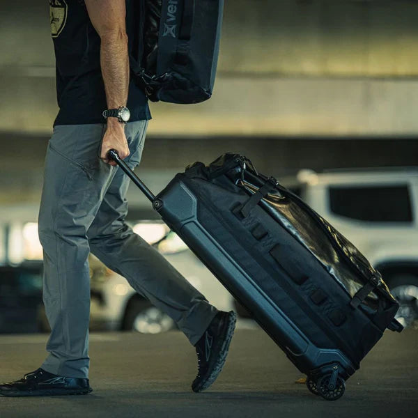 A man pulls a black Vertx Road Less Traveled Duffel Bag across an airport parking lot, utilizing the heavy-duty rolling wheels to transport his gear toward the terminal with ease.