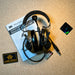 A 3M Peltor ComTac VIII tactical ballistic headset resting on its user manual with two AAA batteries and an Atomic Defense brand patch nearby on a textured surface.