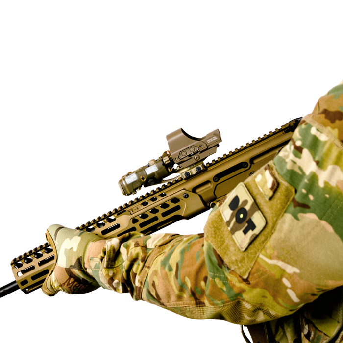 the image shocases the DOT delta optic riser mount in the color FDE or Flat Dark Earth where the item itself is being mounted on top of a weapon and the weapon itself is being held by someone who is wearing a camflauge outfit with a DOT logo patch on its arm. 