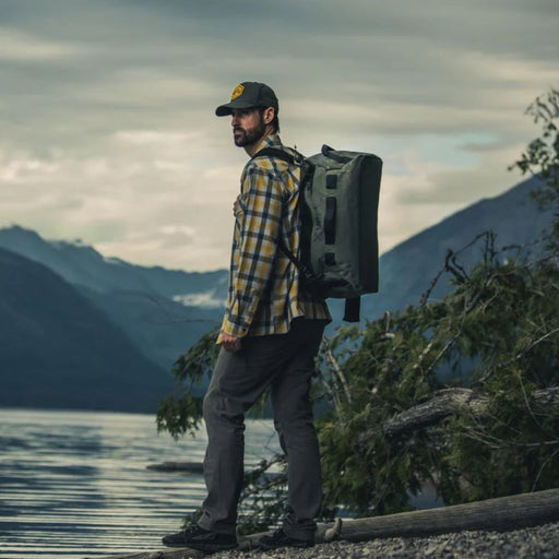 A camper demonstrates the versatility of the Vertx Road Less Traveled 50L Duffel Bag by wearing it in backpack mode while hiking along the edge of a scenic lake .
