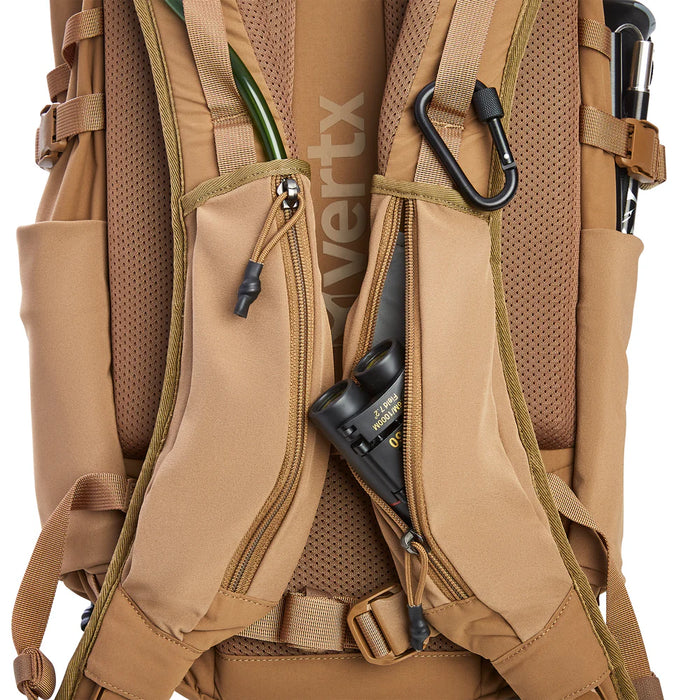 A close-up view of the Vertx Siege 25L tactical backpack shoulder straps shows a small pair of binoculars being inserted into the right easy-access stretch pocket.