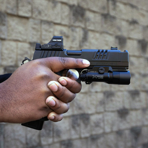 a person holds a handgun with a holosun red dot sight and x300u-b weaponlight, aimed towards the right against a blurred brick wall