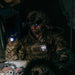 American soldier in tactical gear with night vision goggles holding a bright flashlight in a dark setting.