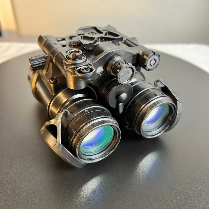 A left-side frontal view of the an/pvs-31d night vision device shows the eyecaps uncovered to reveal the Gen 3 white phosphor intensifier tubes in the foreground. In the background plane, the battery cap, open external power port, and the integrated IR illuminator are clearly visible.