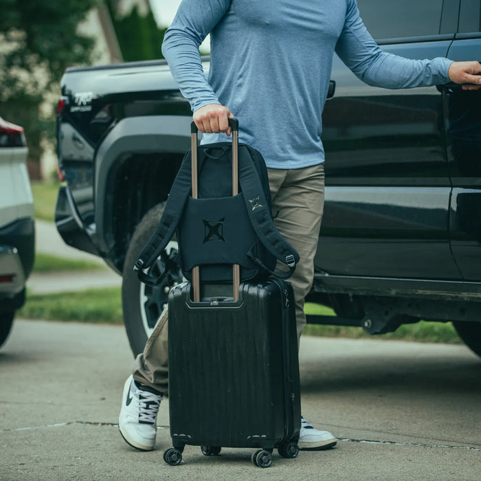 A man preparing to load his pickup truck while carrying the Any Day Backpack, which features a discreet back panel luggage pass-through that easily slides over a rolling suitcase handle for streamlined travel.