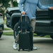 A man preparing to load his pickup truck while carrying the Any Day Backpack, which features a discreet back panel luggage pass-through that easily slides over a rolling suitcase handle for streamlined travel.