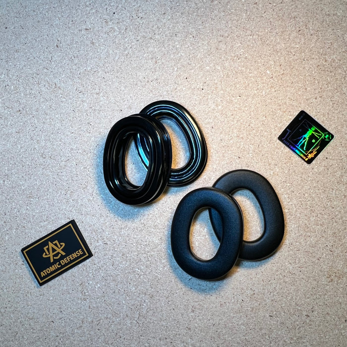 Two pairs of black oval gel earcup replacement seals for a ballistic headset are arranged on a textured tan surface alongside a holographic Atomic Defense sticker.