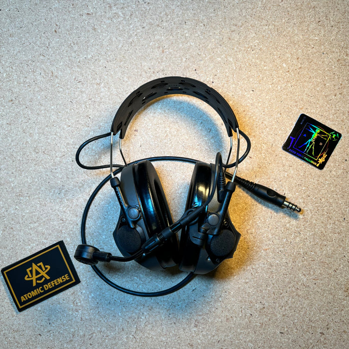 A black 3M Peltor ComTac system engineered for clear battle comms stands upright on a textured surface, featuring a flexible boom microphone and a padded rubberized headband.