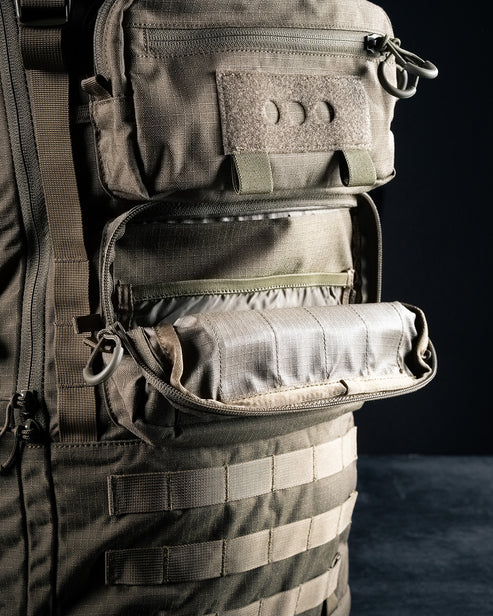A detailed close-up of the lower exterior storage pouch on the green Eberlestock Mission EMOD Ruck, displaying the rugged nylon material and secure zipper closure.