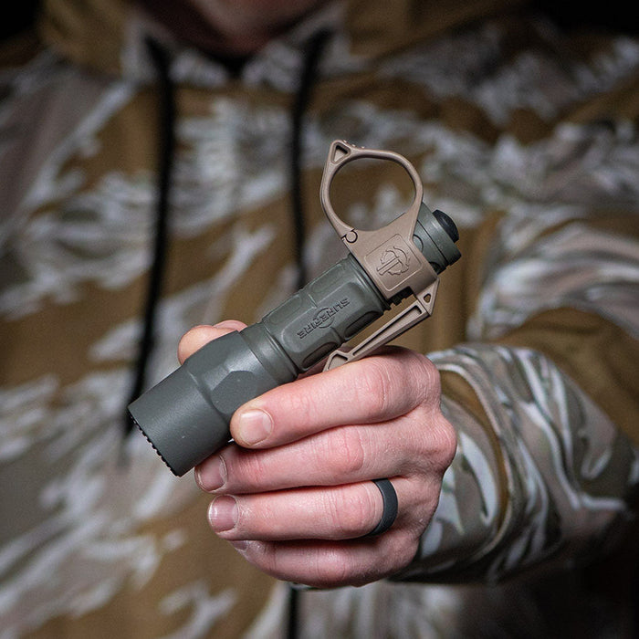 Close-up of a hand in a camouflage jacket holding a green SureFire flashlight with a tan SwitchBack ring.