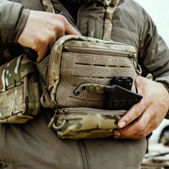 A close-up of a person wearing a camouflage chest rig, actively drawing a black compact pistol from the discreet rear compartment, with a knife and a small drop-down molle pouch attached to the front.