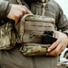 A close-up of a person wearing a camouflage chest rig, actively drawing a black compact pistol from the discreet rear compartment, with a knife and a small drop-down molle pouch attached to the front.