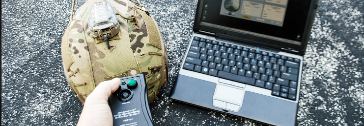 A first-person view showing a hand pointing the CORE Survival HEL-STAR PIM remote at a helmet-mounted beacon, with a laptop displaying the programming software resting on the pavement nearby.