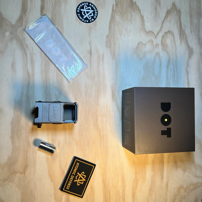 the dot ehs 1 holographic sight presented with its battery pack and box, along with atomic defense patches on each side of it placed on a wooden table