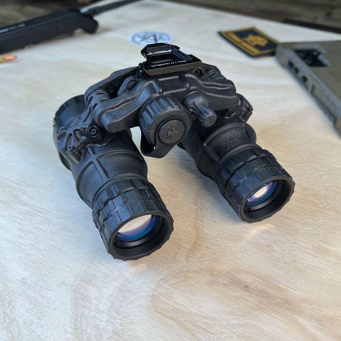 This frontal left angled view displays the DTNVS-MG – (Dual Tube Night Vision System, Manual Gain), showcasing the rugged dual-tube housing and the precision manual gain control knob used to adjust image brightness in different light levels. The perspective highlights the independent monocular pods that can flip up to automatically shut off and save battery life during missions.