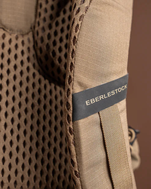 A high-detail close-up of the Eberlestock Brute Scout Pack shoulder strap, featuring the authentic embroidered logo set against the durable ripstop fabric and the edge of the breathable air-mesh padding.