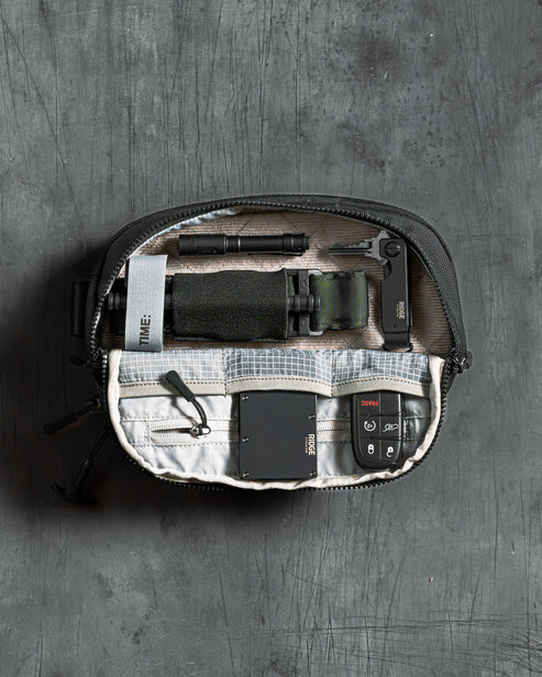 the image showcases the eberlestock fade flex micro in the color black and is placed on a dark surface, where it is also opened up to show how it would look like once filled with items, the velco-in having a knife securely placed, and with the compartments being filled with key remotes or tools.