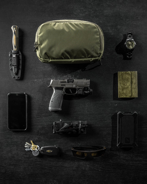 the image shocases the eberlestock fade flex micro in the variant olive where the product is placed on top of a dark wooden table with a lot of items surrounding it as if presenting that these are all the items that will fit inside the product, which is a knife, phone, watch, pistol, magazine, shades, wallet and keys.