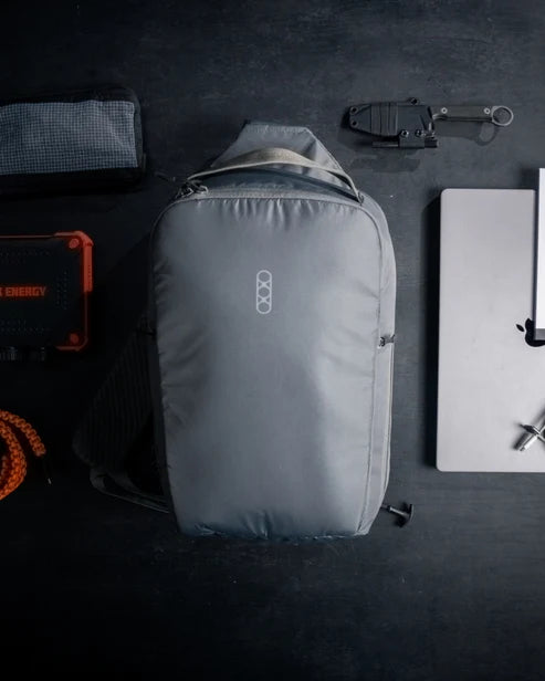 Flat lay of the Eberlestock Fade Sling XL in black surrounded by everyday carry gear, including a portable power bank, folding knife, laptop, and paracord, illustrating a full EDC loadout.