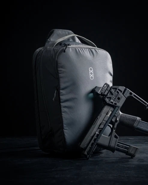 Eberlestock Fade Sling XL in black standing upright on a dark surface beside a disassembled pistol, highlighting the pack's concealed carry purpose.