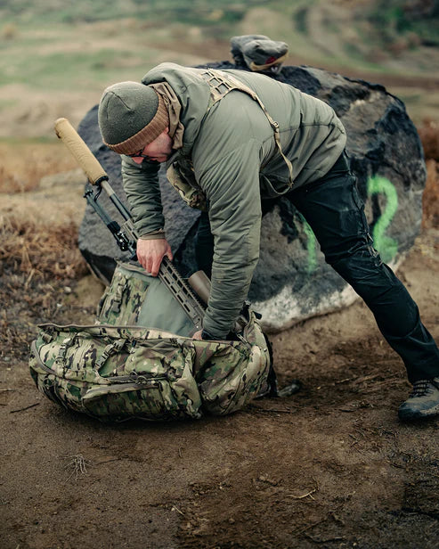 A frontal shot of the Eberlestock Spectre Pack in Multicam camouflage, positioned at a shooting range alongside a Seekins Precision rifle to demonstrate its compatibility with high-end precision weapon systems.