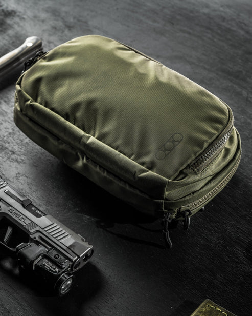 an image of the eberlestock fade flex micro in the olive variant and is being placed on a dark table beside a small pistol. 