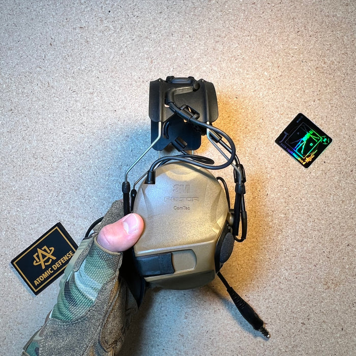A gloved hand holds a folded coyote brown 3M Peltor ComTac headset, displaying the exterior earcup branding and the integrated downlead cable used to establish secure battle comms in tactical environments.
