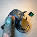 A gloved hand securely grips a folded coyote brown 3M Peltor ComTac headset, displaying the flexible rubberized headband bent over the earcups for compact tactical comms storage.