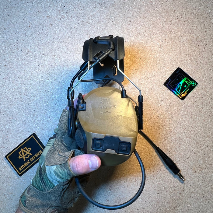 A close-up view of a gloved hand holding a folded coyote brown 3M Peltor ComTac tactical headset, highlighting the exterior earcup's integrated volume control buttons and communication downlead cable.