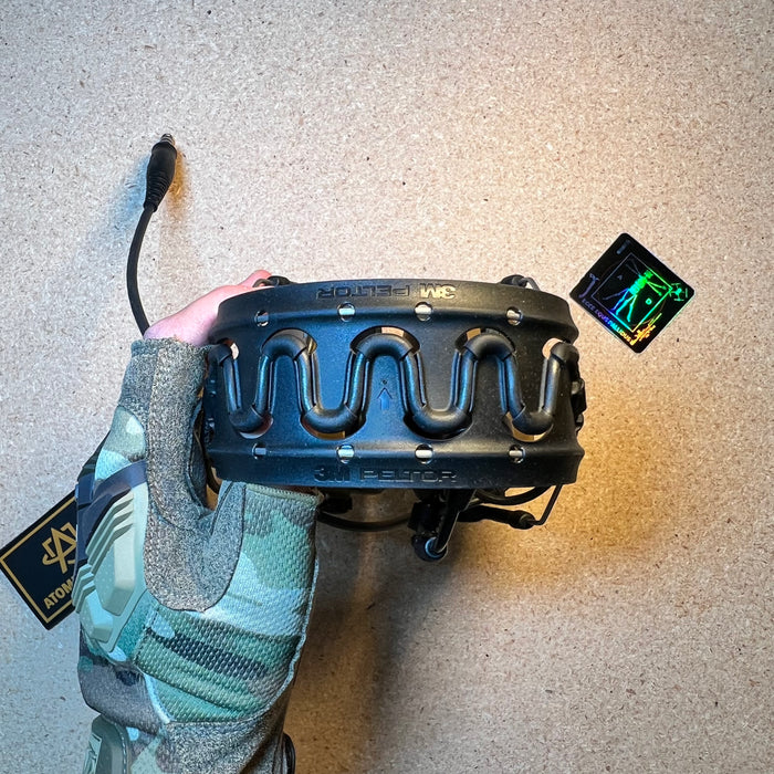 A gloved hand displays the top of a black rubberized headband belonging to a Peltor ComTac VIII tactical headset, highlighting the flexible, ventilated cutout design and 3M branding against a textured tan surface.