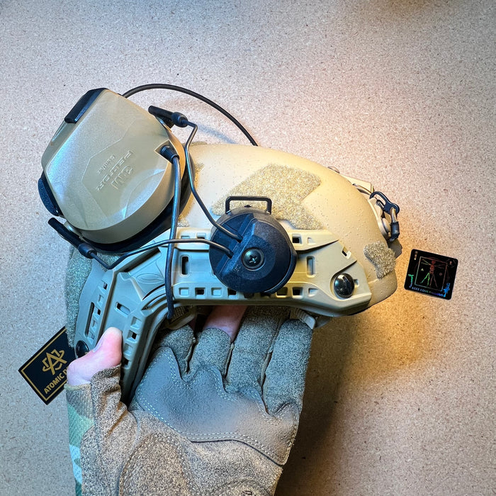 A gloved hand holds a tan high-cut ballistic helmet equipped with an olive drab green rail-mounted tactical headset, showing the exterior earcups and communication wiring.