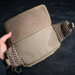 A person's hand holding open the rear section of an olive green pack, revealing a wide hook-and-loop area designed to securely attach a drop-down molle panel or other accessories.