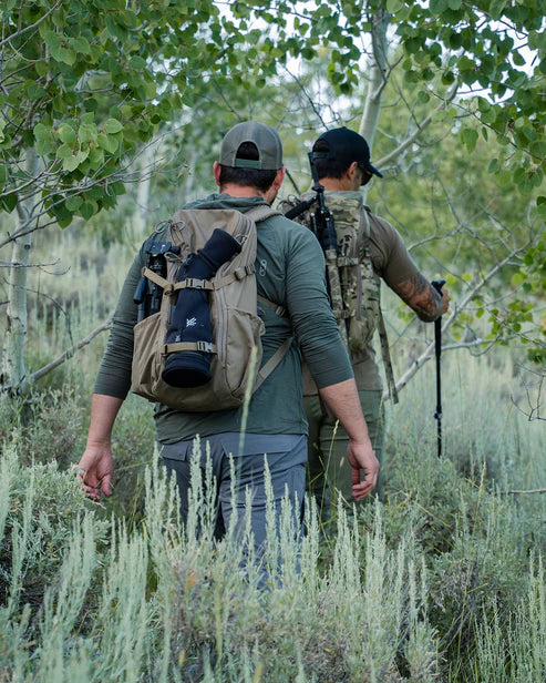 Two hikers navigate a ridgeline wearing the Eberlestock Brute Scout 30L Hunting Pack, with one operator using the Multicam variant and the other the Dry Earth version, demonstrating how the pack's low-profile design stays tight to the body during movement.