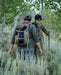 Two hikers navigate a ridgeline wearing the Eberlestock Brute Scout 30L Hunting Pack, with one operator using the Multicam variant and the other the Dry Earth version, demonstrating how the pack's low-profile design stays tight to the body during movement.