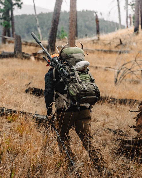 A hunter trekking through the backcountry wearing the Eberlestock Mainframe II Pack Frame in the Mountain colorway, fully loaded with a Vapor 2500 bag, a secured rifle, and a harvested trophy head on the meat shelf.