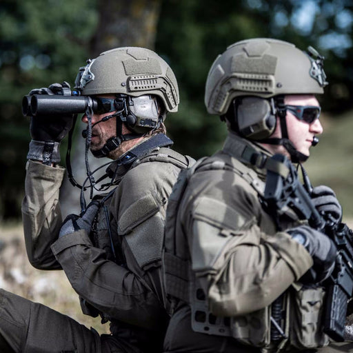two members of armed forces wearing tactical helmets with peltor comtac viii headsets are positioned outdoors, with one using binoculars and the other holding a rifle