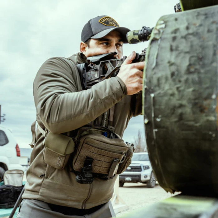 A male shooter wearing a baseball cap and an olive green jacket aims a rifle with a mounted optic, utilizing the hands-free mobility provided by modular tactical chest packs during range training.