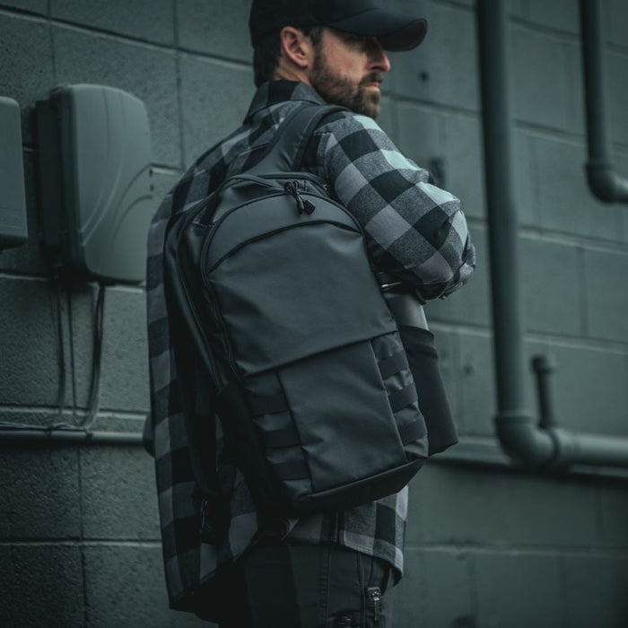 man wearing grey flannel shirt and black cap carries vertx urban ghost backpack on right shoulder