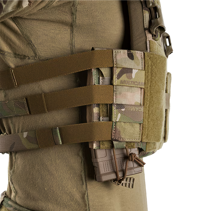 A close-up side view of an operator wearing a tactical vest demonstrating the crye precision low-profile visibility setup. The MultiCam Side Radio Pouch is mounted on the side straps and configured in an inverted orientation, securely holding a brown rifle magazine downwards using the shock cord retention system.