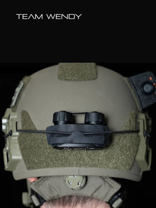 the image shows a product model wearing a ranger green or sage team wendy helmet that has the nocturn industries mini bat pack in the black version that is attached on the helmet using the velcro backing of the battery pack and secured with the bungee/shock cored through the built in retention loops and is anchored to the helmet's rear ARC rail slots.