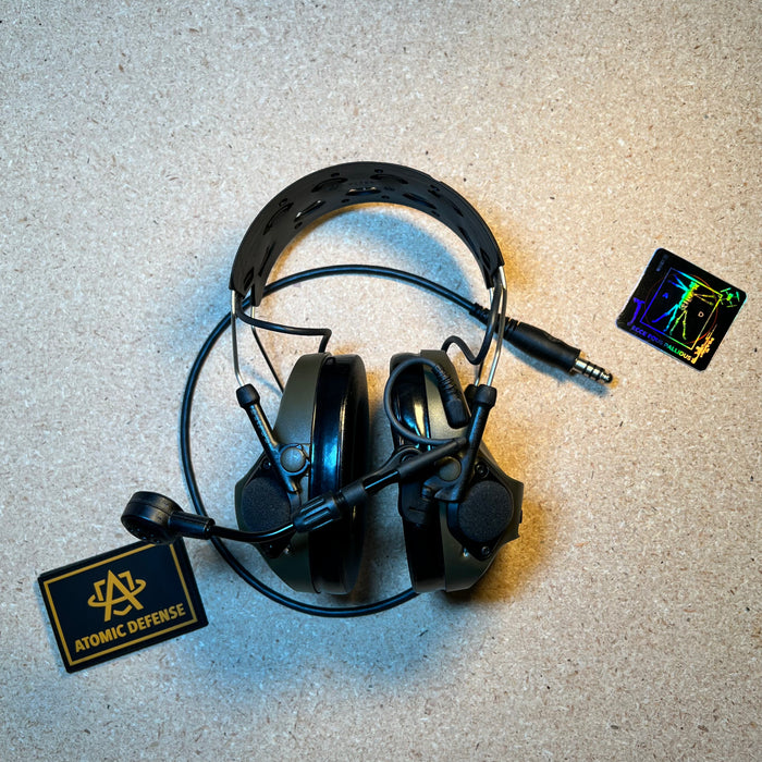 An olive drab green tactical ballistic headset stands upright on a textured surface, displaying its flexible boom microphone and adjustable headband next to an Atomic Defense patch.