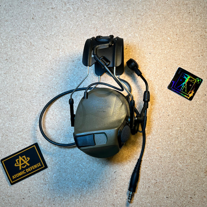 An olive drab green 3M Peltor ComTac tactical headset with helmet rail mounting brackets and communication downlead cable rests on a textured tan background.