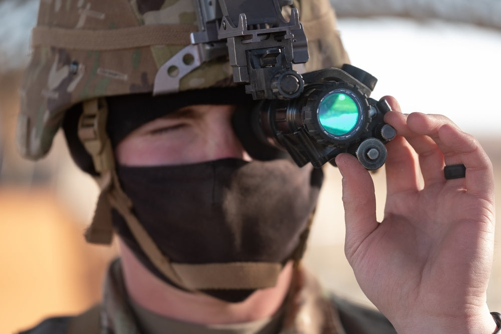 A tactical operator's hand reaches up to adjust the manual gain knob on a pvs 14 nvg unit, which is securely mounted to a helmet using a Wilcox J-arm and a Wilcox G24 mount.