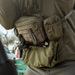 An outdoor operator wearing an olive green chest rig equipped with a folding knife clipped to the front, a secondary tactical pouch attached to the side, and a drop-down handwarmer secured below.
