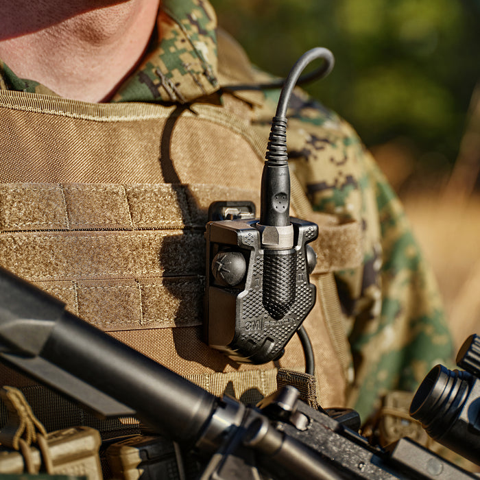 A close-up shot of the Peltor Dual PTT Adapter securely mounted to a Marine plate carrier, demonstrating how the compact, rugged housing attaches to tactical gear for easy access to dual radio controls during field operations.